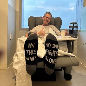 Man sits in a hospital chair with his feet up and on the bottom of his socks says "In this family no one fights alone"