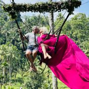 A man and woman in a flowing pink dress sit atop a harness among the jungle trees kissing