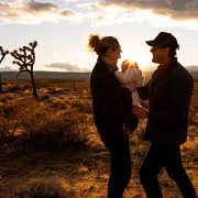 Man and woman stand in a desert holding a baby between them at sunrise.